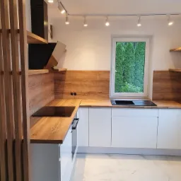 Nowoczesna kuchnia z białymi szafkami, drewnianym blatem i panelem ściennym, widok na zieleń za oknem, drewniana ażurowa ścianka działowa po lewej stronie.