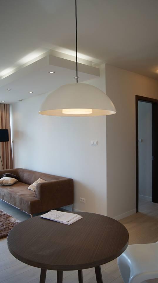 Nowoczesny salon z brązową sofą, okrągłym stolikiem z dokumentami i wiszącą lampą kopułową z podświetleniem LED w suficie podwieszanym.