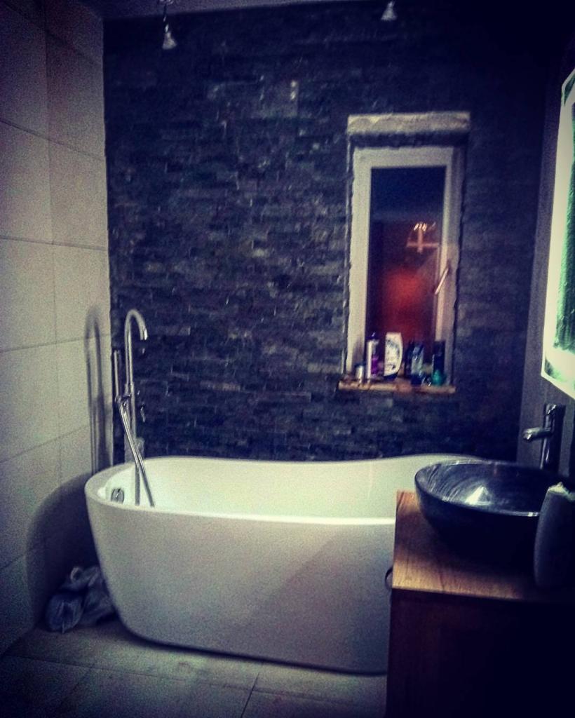 Nowoczesna łazienka z wolnostojącą białą wanną, czarną kamienną ścianą i umywalką w kształcie misy na drewnianej szafce, oświetlona punktowymi lampami sufitowymi i światłem z okna.