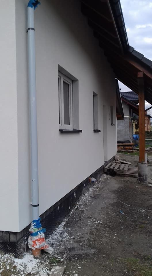 Świeżo otynkowana, biała elewacja domu jednorodzinnego z widocznym systemem rynnowym zabezpieczonym folią, czarną izolacją fundamentów i pozostałościami zaprawy na gruncie.