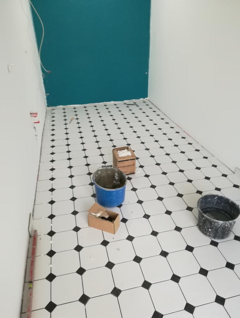 Układanie czarno-białej podłogi w łazience z turkusową ścianą, widoczne wiaderka, poziomica i pudełko z fugą.