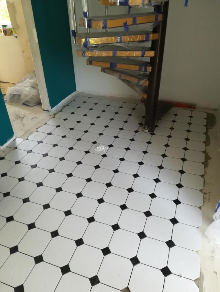 Ułożona podłoga z białych, ośmiokątnych płytek z czarnymi kwadratami w narożach, widoczne zabezpieczone folią schody spiralne i fragment pomalowanej na turkusowo ściany.