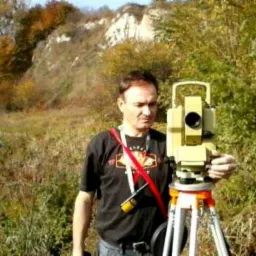 Mężczyzna w czarnej koszulce obsługuje żółty teodolit na trójnogu, w tle porośnięte wzgórze i jesienne drzewa.