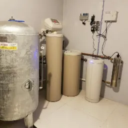 Zestaw urządzeń do uzdatniania wody w kotłowni, widoczny hydrofor z naklejką informacyjną, filtry kolumnowe oraz lampa UV, instalacja rurowa na ścianie.