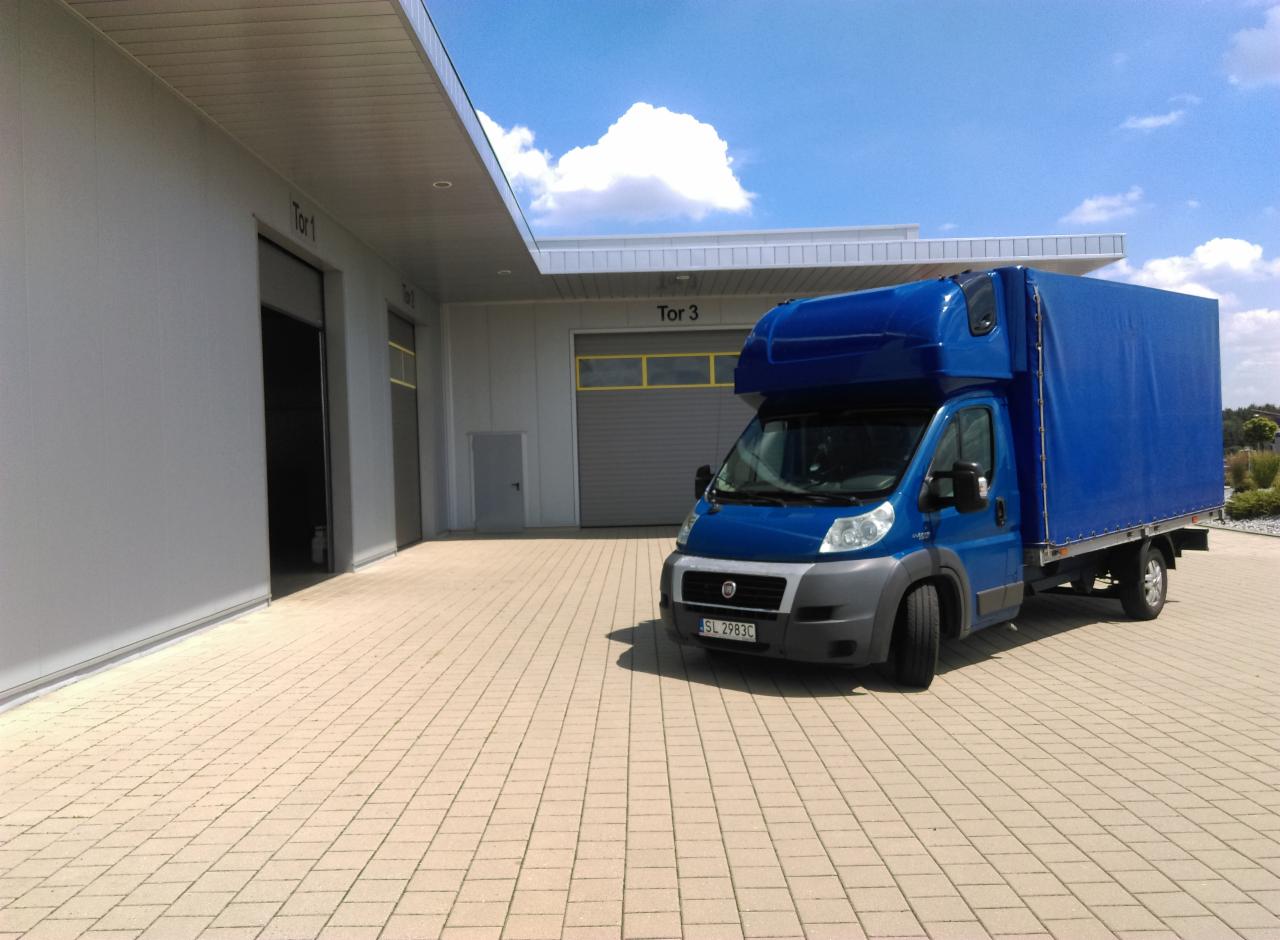 Niebieski samochód dostawczy z plandeką zaparkowany przed budynkiem magazynowym z bramami 'Tor 1' i 'Tor 3' w słoneczny dzień.