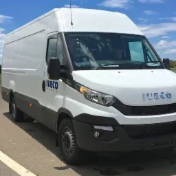 Biały samochód dostawczy marki Iveco na asfalcie z pomarańczowymi pachołkami w tle, widok z przodu.