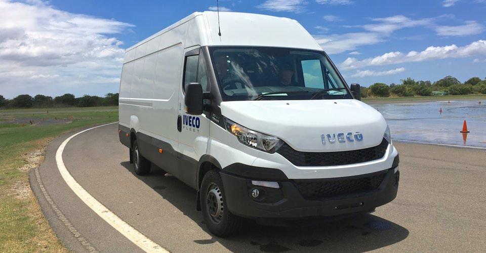 Biały samochód dostawczy marki Iveco na asfalcie z pomarańczowymi pachołkami w tle, widok z przodu.