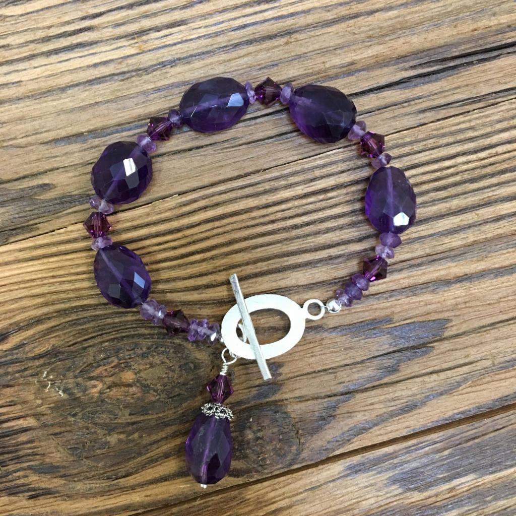 Bransoletka z fasetowanych ametystów i srebrnym zapięciem typu toggle, leżąca na drewnianym tle z widocznym usłojeniem.