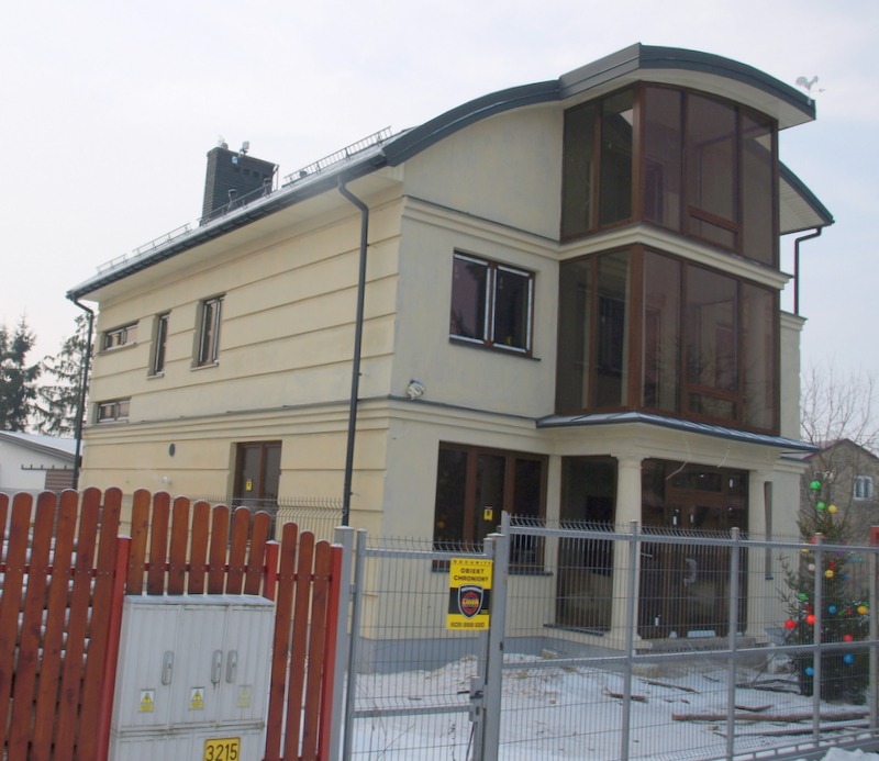 Dwupiętrowy dom z jasną elewacją, przeszkloną dobudówką i ciemną stolarką okienną, widoczny zza metalowego ogrodzenia i drewnianego płotu, z choinką w rogu działki.