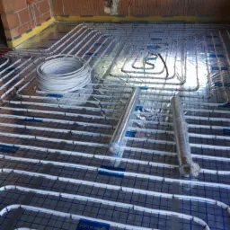 Rozłożona instalacja ogrzewania podłogowego na folii z siatką, widoczne rury w zwoju i rolki folii izolacyjnej, surowe ściany z cegły.