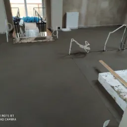 Świeżo wylana, gładka, szara posadzka w surowym wnętrzu budynku. Widoczne wystające rurki ogrzewania podłogowego i przewody elektryczne. W tle schody i biała szafka.