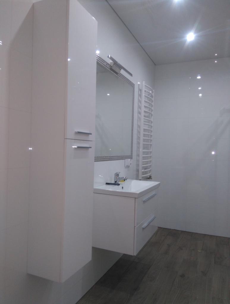 Nowoczesna łazienka z białymi, błyszczącymi płytkami, drewnopodobną podłogą, wiszącą szafką z umywalką i wysoką szafką boczną oraz grzejnikiem drabinkowym.