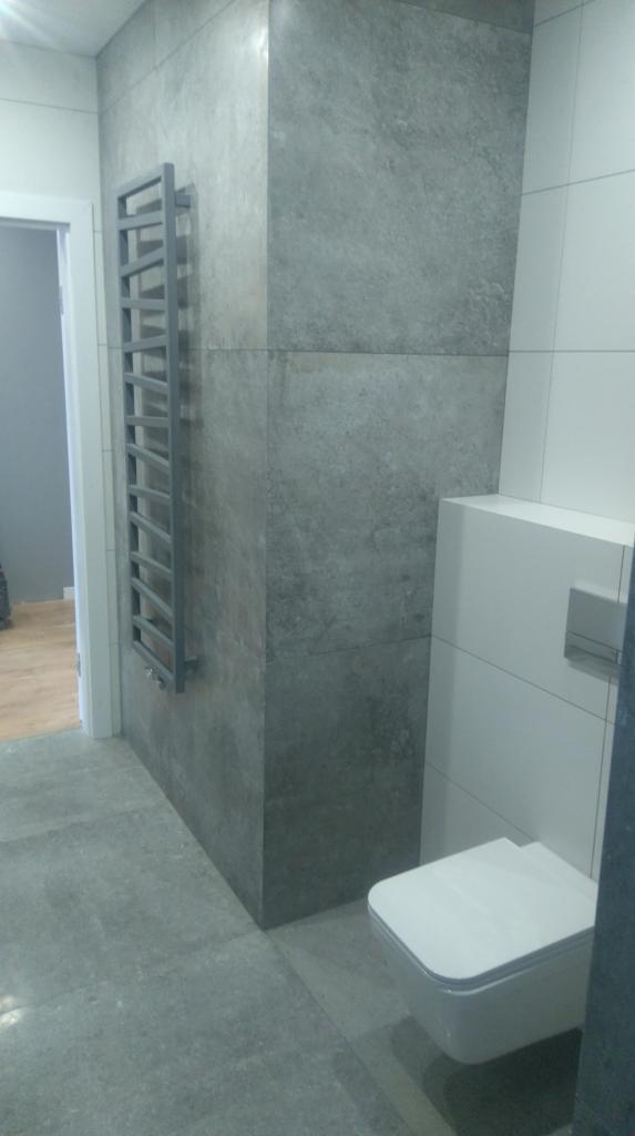 Nowoczesna łazienka z szarymi, wielkoformatowymi płytkami imitującymi beton, grzejnikiem drabinkowym i podwieszaną toaletą.