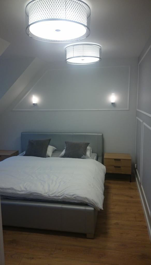 Nowoczesna sypialnia na poddaszu z szarą tapetą, listwami dekoracyjnymi, dwoma okrągłymi lampami sufitowymi z metalową siatką i drewnianą podłogą.