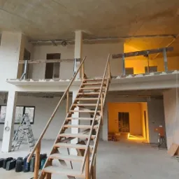 Wnętrze dwupoziomnowego domu w stanie deweloperskim z drewnianymi schodami, częściowo otynkowanymi ścianami i widoczną instalacją elektryczną.