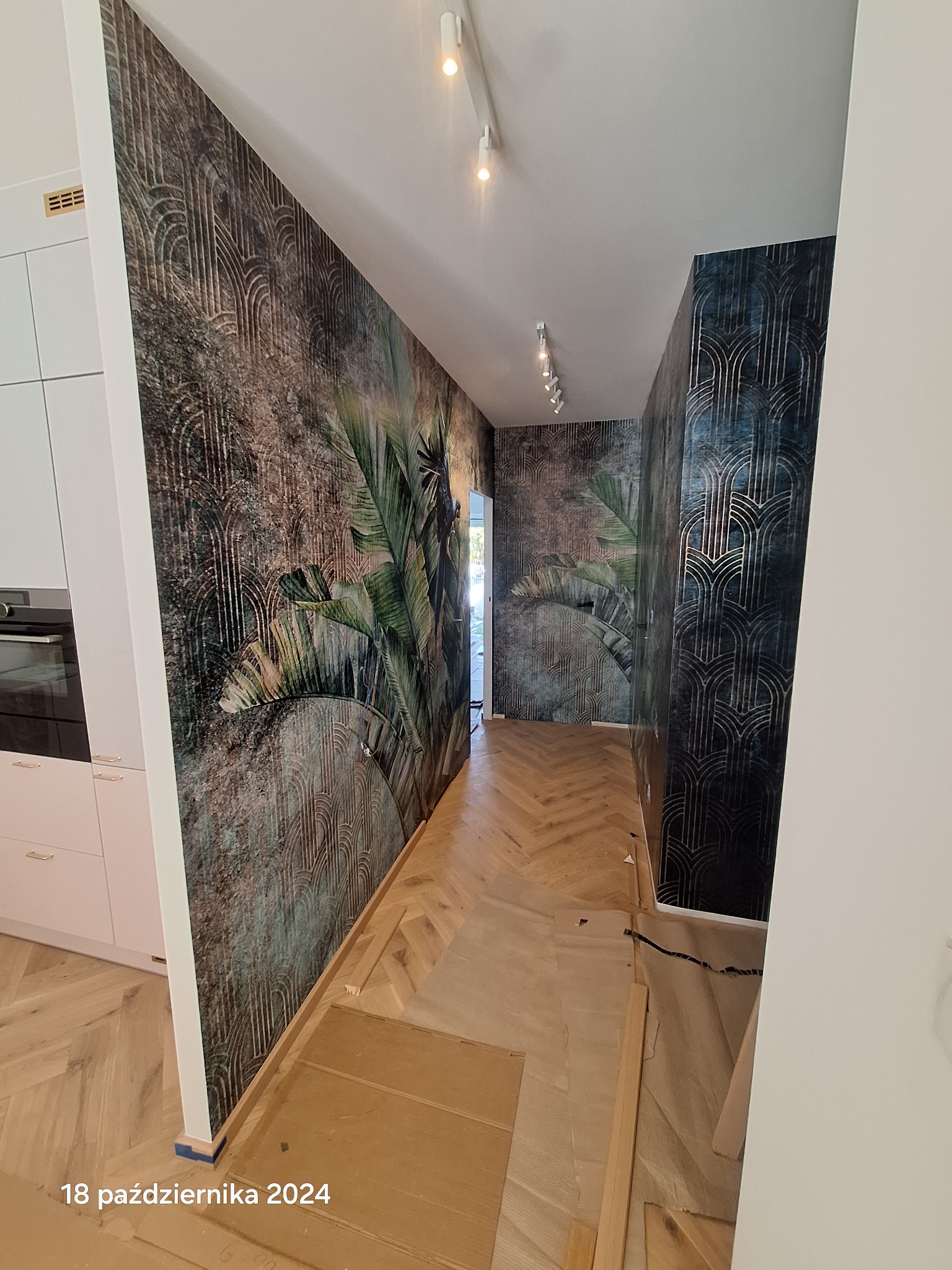 Korytarz po metamorfozie: wzorzyste tapety w stylu art deco i motywy roślinne, parkiet w jodełkę, oświetlenie punktowe. Widoczne niedokończone prace przy podłodze.