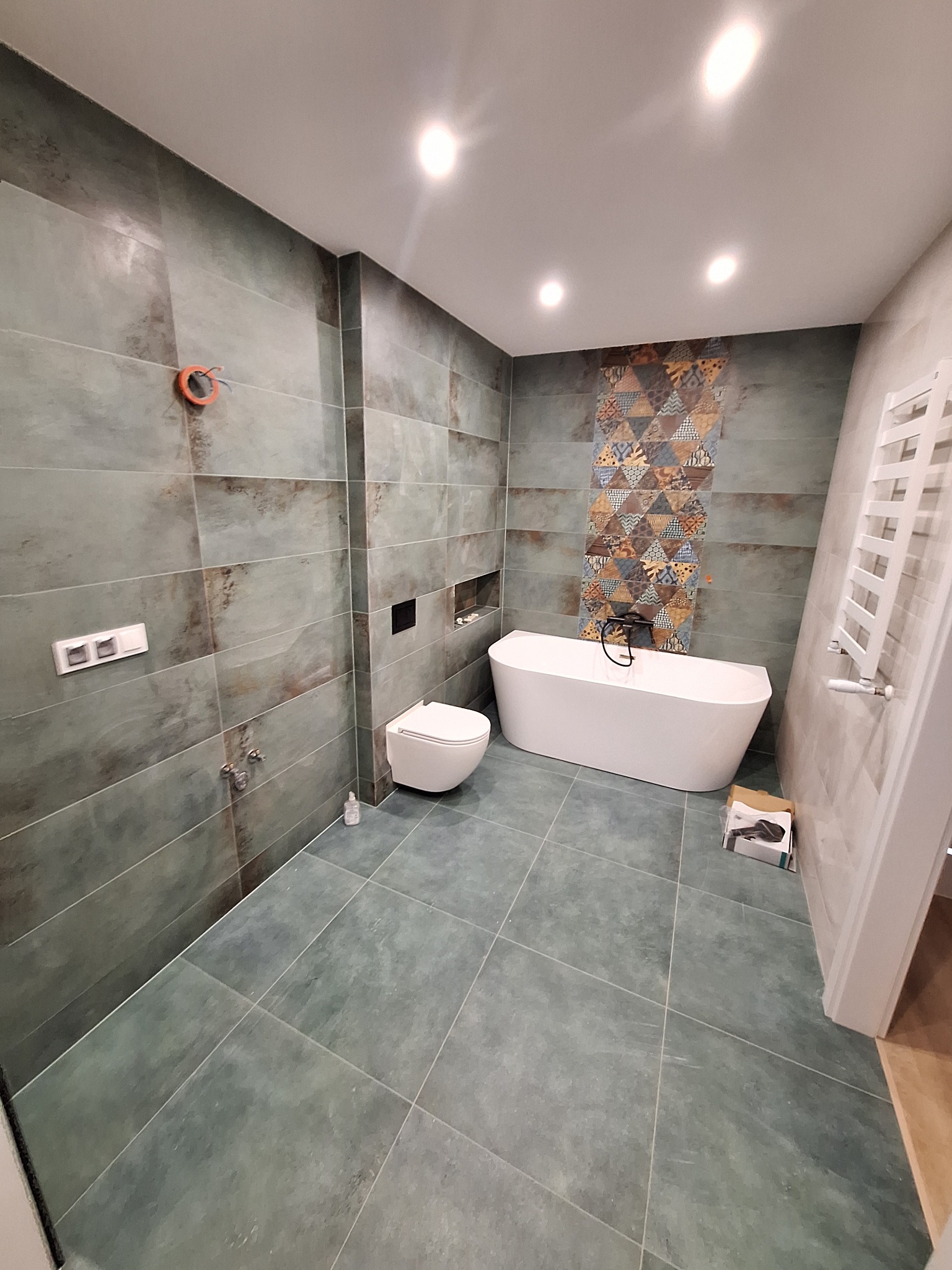 Nowoczesna łazienka z wanną wolnostojącą, toaletą podwieszaną i płytkami w odcieniach szarości z dekoracyjnym pasem mozaiki. Grzejnik drabinkowy i minimalistyczne oświetlenie.