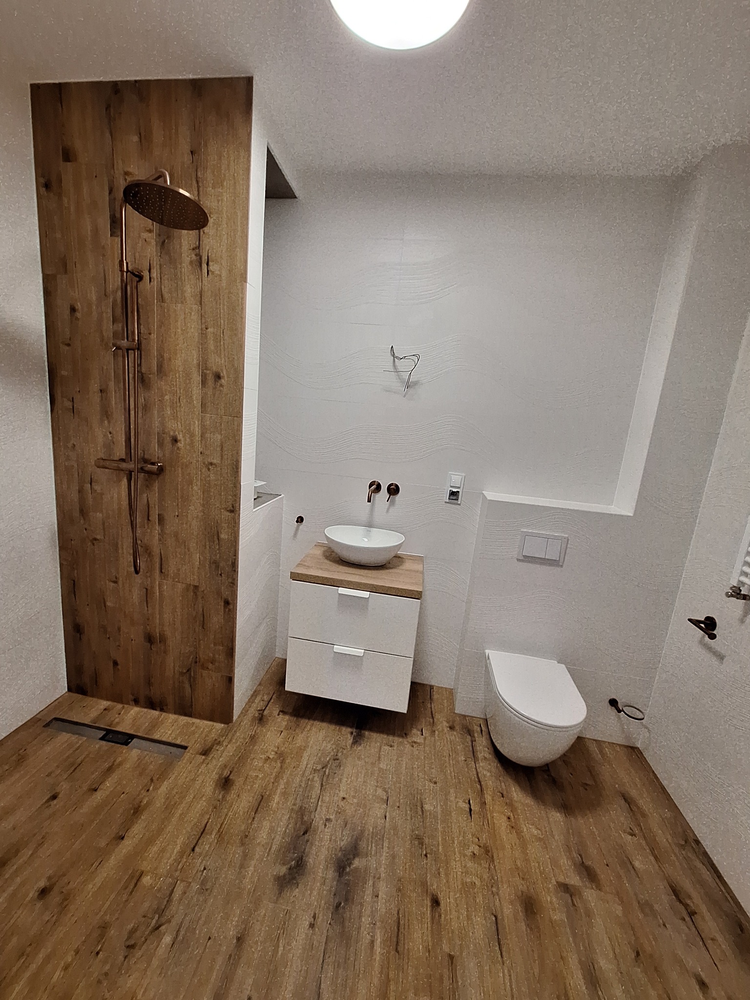 Nowoczesna łazienka z drewnopodobną podłogą i ścianą prysznicową, białą armaturą i miedzianą baterią. Minimalistyczny design z akcentami naturalnego drewna.