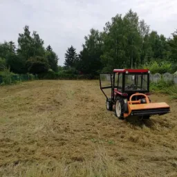 Mały traktor z kosiarką rotacyjną na polu po skoszeniu trawy, widoczne drzewa i dom w tle.
