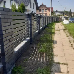 Nowoczesne ogrodzenie z grafitowej cegły klinkierowej i poziomych paneli, widok z boku na tle domów jednorodzinnych i chodnika.