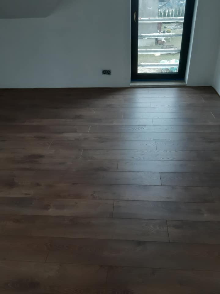 Ukończona podłoga z paneli w odcieniach ciemnego brązu, widok na białą ścianę z gniazdkami elektrycznymi i okno dachowe z czarną ramą.