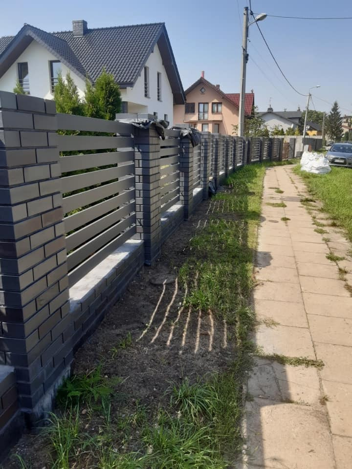 Nowoczesne ogrodzenie z grafitowej cegły klinkierowej i poziomych paneli, widok z boku na tle domów jednorodzinnych i chodnika.