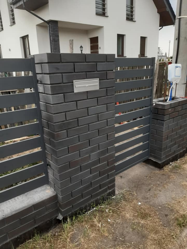 Murowane słupki z szarej cegły klinkierowej, z wbudowaną skrzynką na listy i przęsłami metalowego ogrodzenia, na tle elewacji domu.