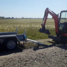 Pomarańczowa mini koparka Kubota KX016-4 z opuszczoną rampą na przyczepie dwuosiowej, na szutrowej drodze, w tle pole zboża pod błękitnym niebem.