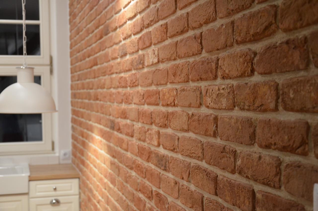 Dekoracyjna ściana z czerwonej cegły w kuchni, widok z boku, z elementami białej zabudowy kuchennej i lampą wiszącą w tle.
