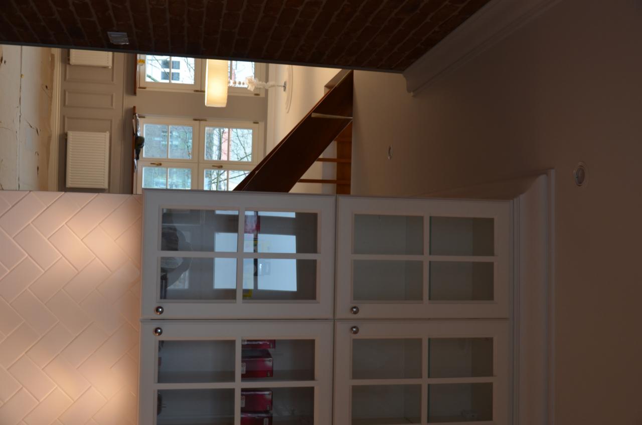 Wnętrze w trakcie remontu: biała ściana z ułożonymi płytkami w jodełkę, białe szafki z przeszklonymi drzwiczkami, widoczny fragment schodów i okno z widokiem na zewnątrz.