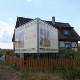 Billboard reklamujący 'Wioskę Domów Modelowych DANMAR domy drewniane' na tle domu z dachem krytym strzechą i drewnianym ogrodzeniem.