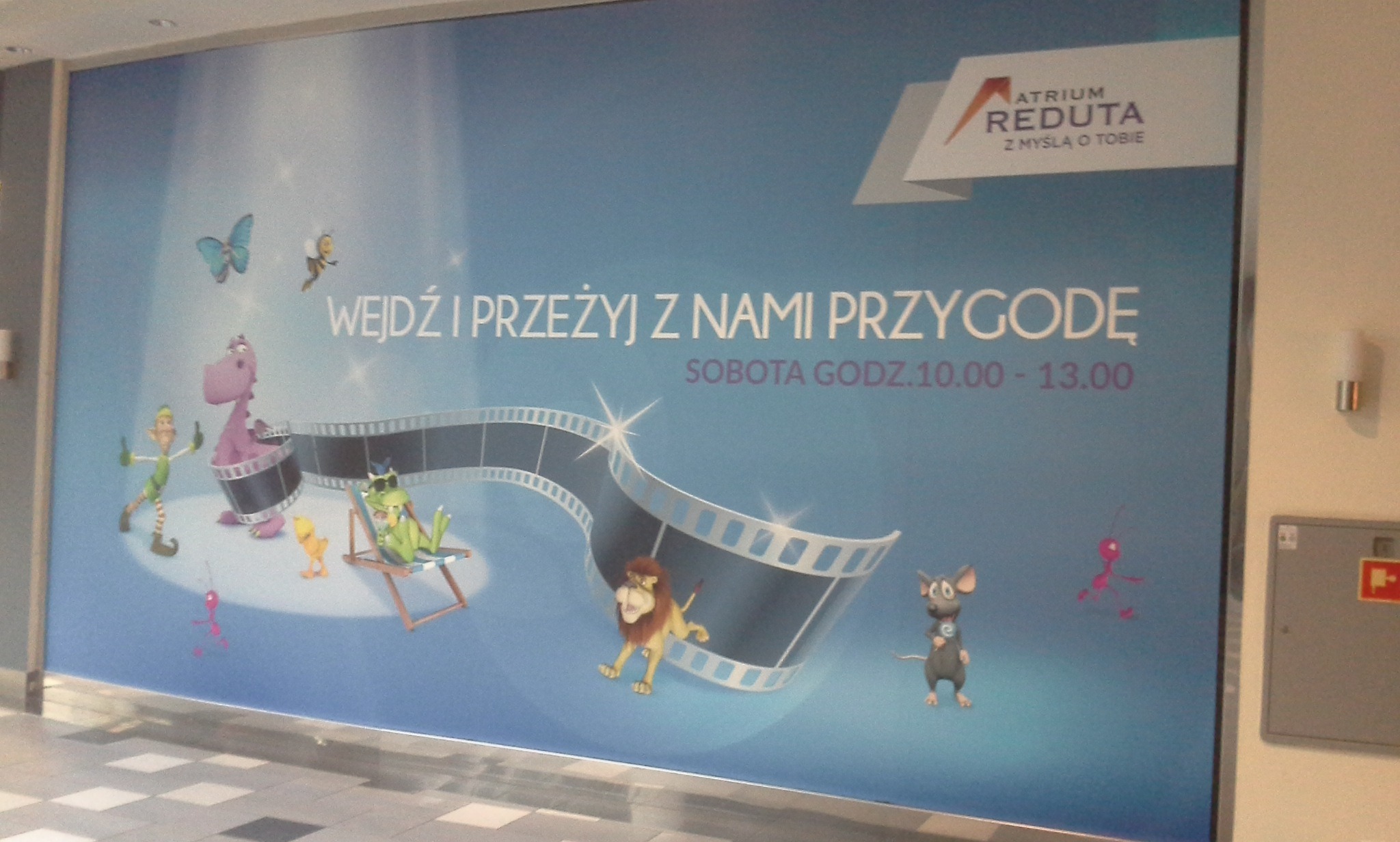 Wielkoformatowy druk z animowanymi postaciami i napisem 'Wejdź i przeżyj z nami przygodę' w centrum handlowym Atrium Reduta.