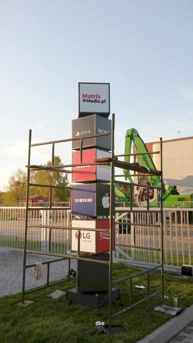 Totem reklamowy złożony z kostek z logotypami różnych firm (Matrix Media, Playstation, Sony, Micro, Karcher, Samsung, Apple, LG, Brita) na tle budowy i zielonego dźwigu