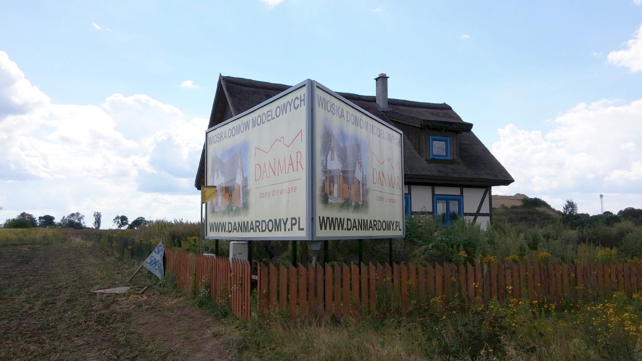 Billboard reklamujący 'Wioskę Domów Modelowych DANMAR domy drewniane' na tle domu z dachem krytym strzechą i drewnianym ogrodzeniem.