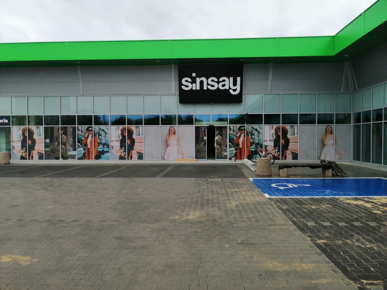 Zewnętrzna reklama na szklanej fasadzie sklepu Sinsay, prezentująca fotorealistyczne wydruki w oknach z wizerunkami modelek w różnych stylizacjach, widoczne z parkingu.