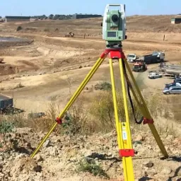 Żółty teodolit na statywie na tle placu budowy z wykopami, maszynami budowlanymi i zaparkowanymi samochodami w słoneczny dzień.