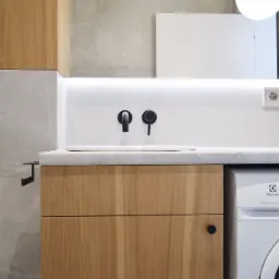 Nowoczesna łazienka z drewnianymi szafkami, umywalką z czarną armaturą, pralką Electrolux i szarą marmurową okładziną ścienną.