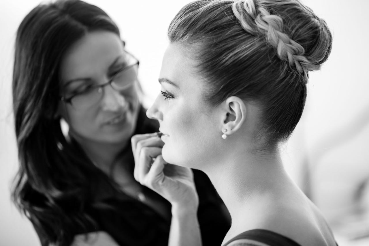 Czarno-białe zdjęcie kobiety z upiętymi włosami w warkocz podczas wykonywania makijażu przez wizażystkę w okularach.