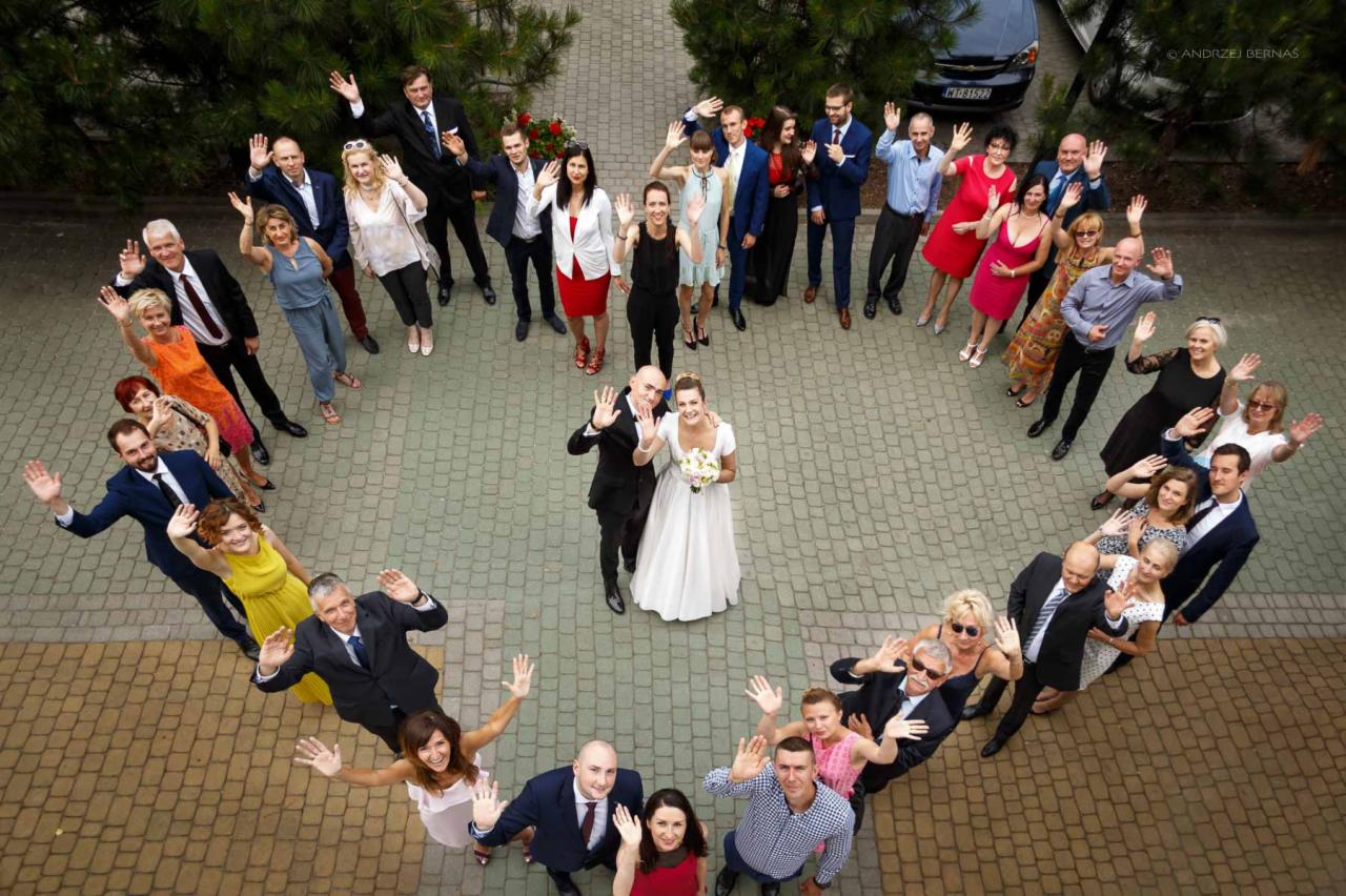 Para młoda otoczona gośćmi weselnymi ustawionymi w kształt serca, wszyscy machają do obiektywu, ujęcie z góry na brukowanym placu.