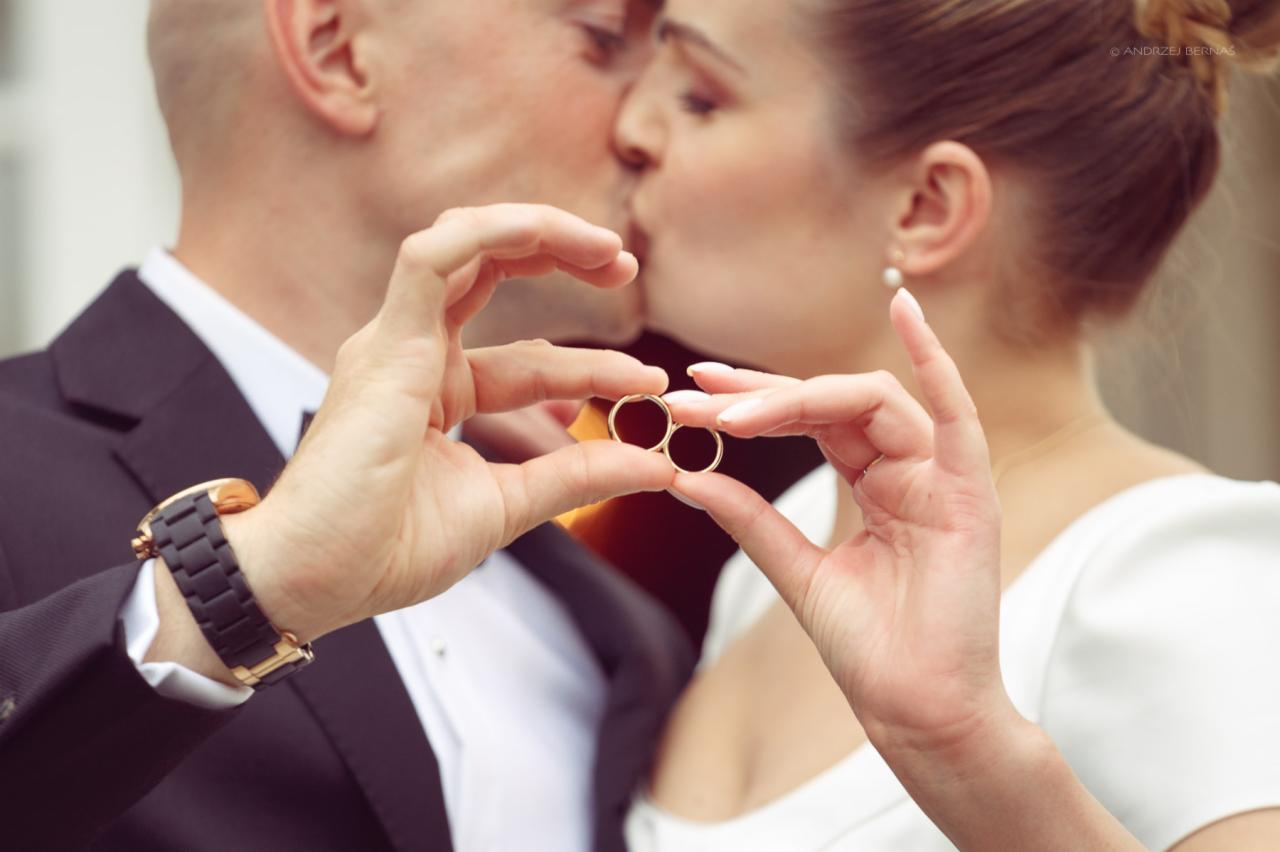 Pocałunek pary młodej w tle, na pierwszym planie dłonie trzymające obrączki ślubne, męska dłoń z zegarkiem na czarnym pasku.