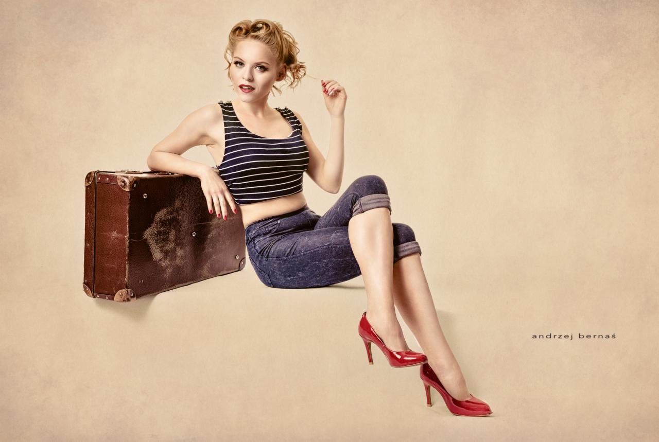 Kobieta w stylu pin-up siedzi na starej walizce, ubrana w paski top i dżinsy, czerwone szpilki, z lokami we włosach i czerwonymi ustami.