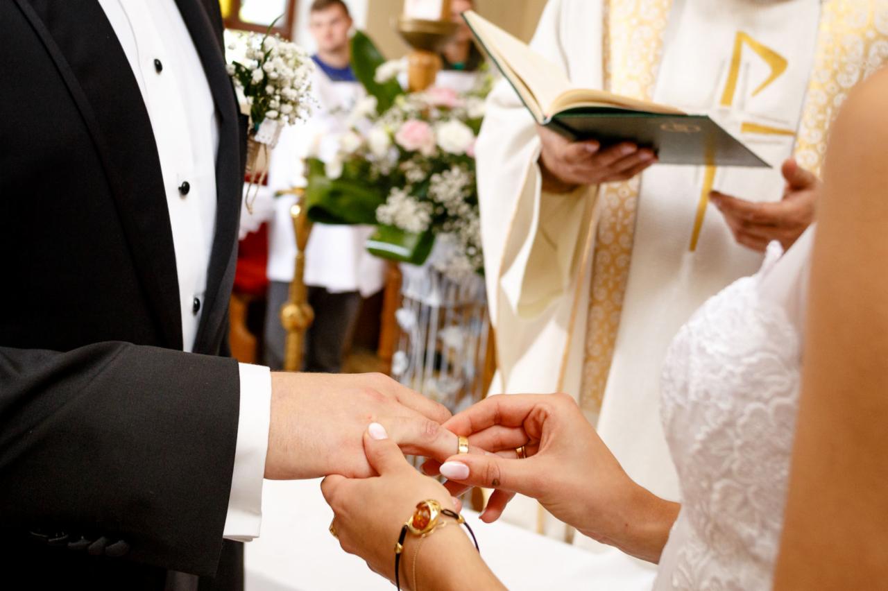 Moment zakładania obrączki podczas ceremonii ślubnej, widoczny fragment sukni panny młodej, garnitur pana młodego oraz ręce księdza z otwartą księgą w tle.