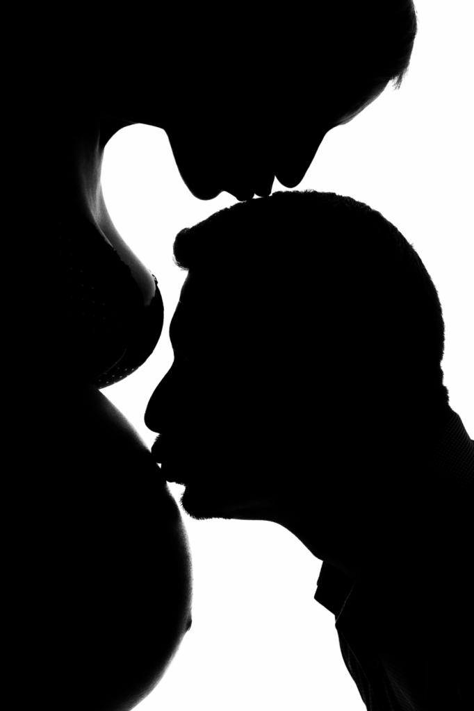 Sylwetki kobiety w ciąży i mężczyzny, on całuje jej brzuch, ona dotyka czołem jego głowy, czarno-biała fotografia.