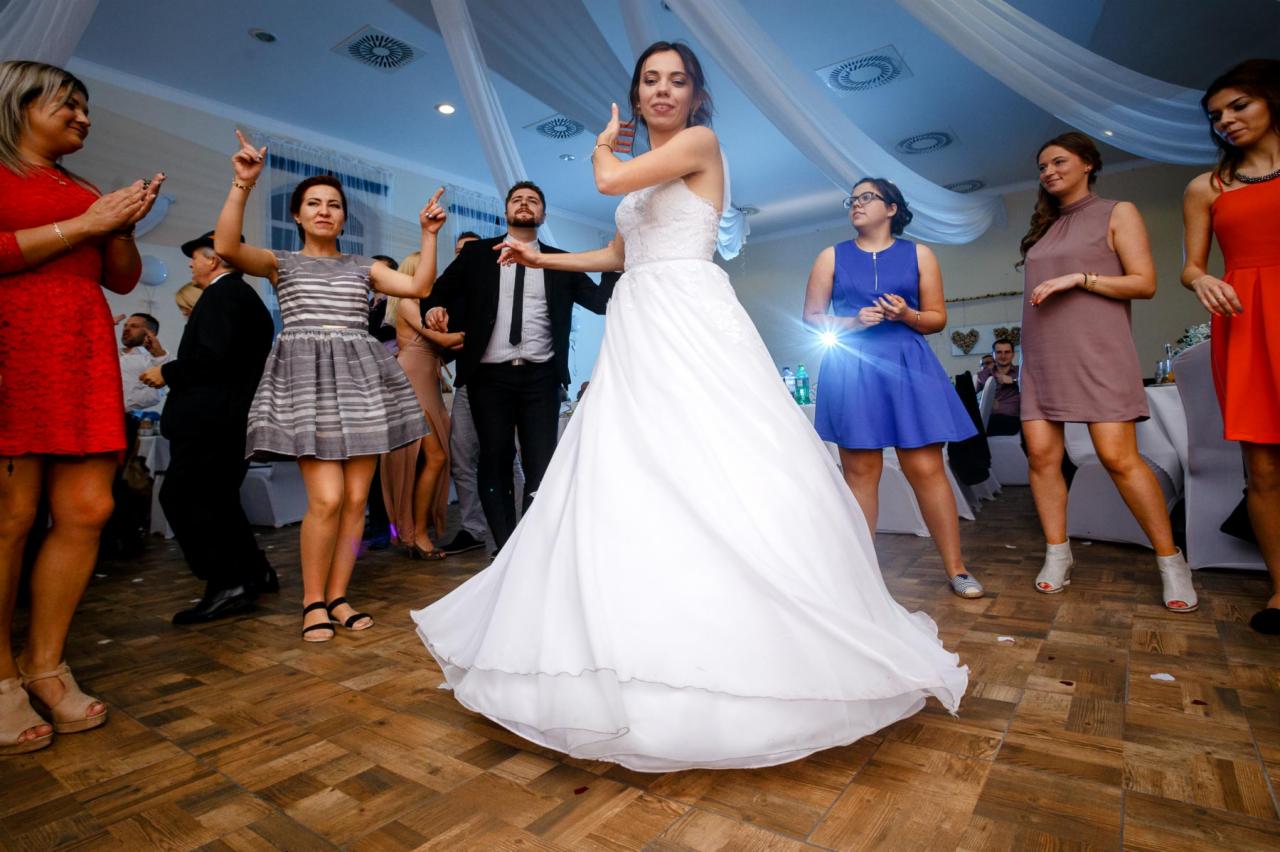 Panna młoda w białej sukni tanecznej, otoczona gośćmi weselnymi na parkiecie z drewnianych paneli, oświetlonym od góry dekoracyjnymi tkaninami.