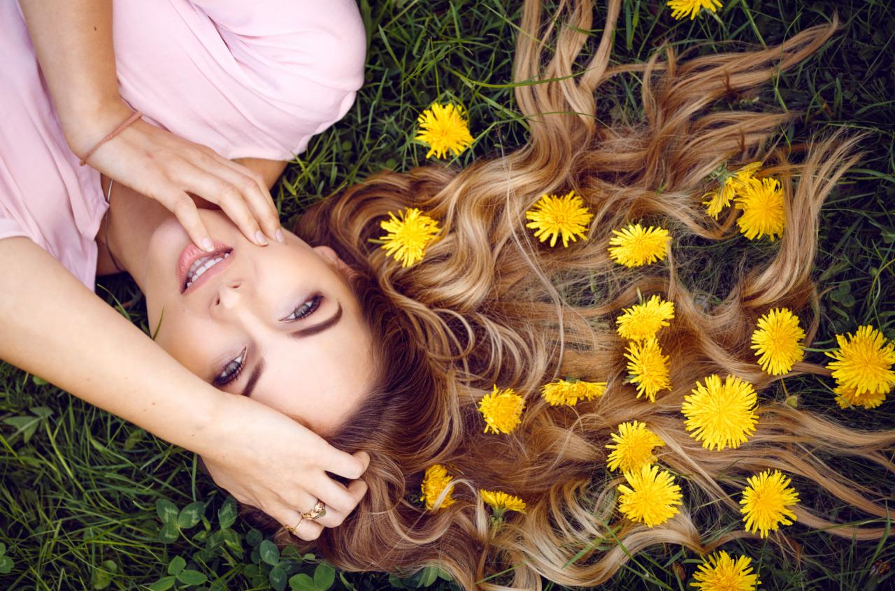 Kobieta leżąca na trawie z rozpuszczonymi włosami ozdobionymi żółtymi kwiatami, delikatny uśmiech, pierścionek na palcu.