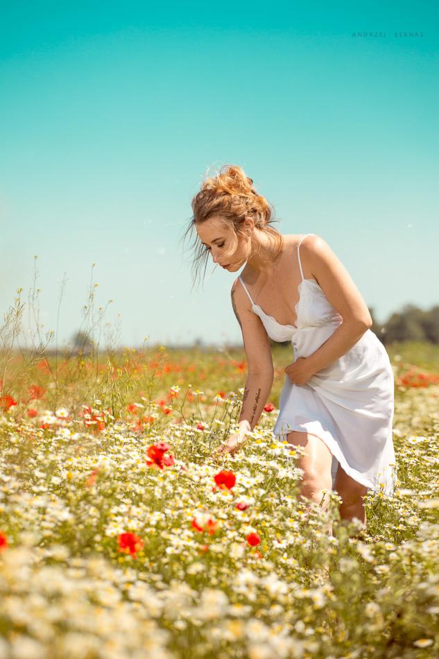 Kobieta w białej sukience na łące pełnej kwitnących rumianków i maków, z tatuażem na ramieniu, w letnim słońcu.