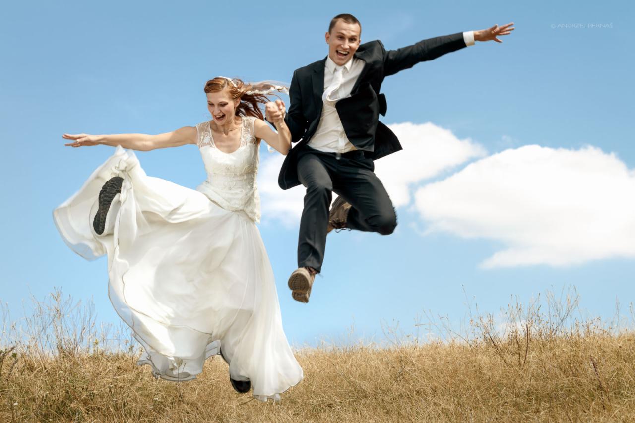 Radosna para młoda, trzymając się za ręce, skacze na tle błękitnego nieba z chmurami i pola z wyschniętą trawą; widoczna podeszwa buta panny młodej.