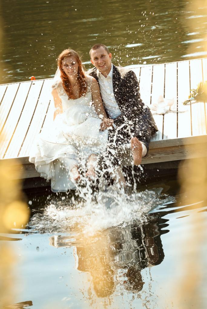 Uśmiechnięta para młoda siedzi na drewnianym pomoście, zanurzając stopy w wodzie, tworząc efektowne rozbryzgi.
