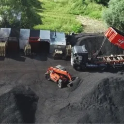 Widok z g&oacute;ry na teren składu z hałdami czarnego węgla, ciężar&oacute;wką z podniesioną skrzynią wyładowującą węgiel oraz pomarańczową ładowarką i zadaszonymi magazynami w tle.