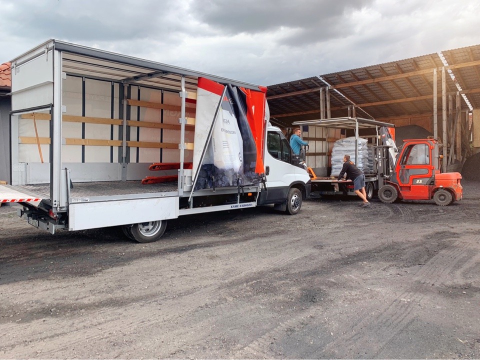 Rozładunek worków z ekogroszkiem z użyciem wózka widłowego i samochodu dostawczego na terenie składu opału, widoczne ślady węgla na podłożu.